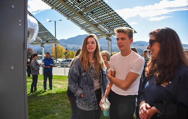 Two high school students and their teachers look at an electric meter while standing under solar panels.